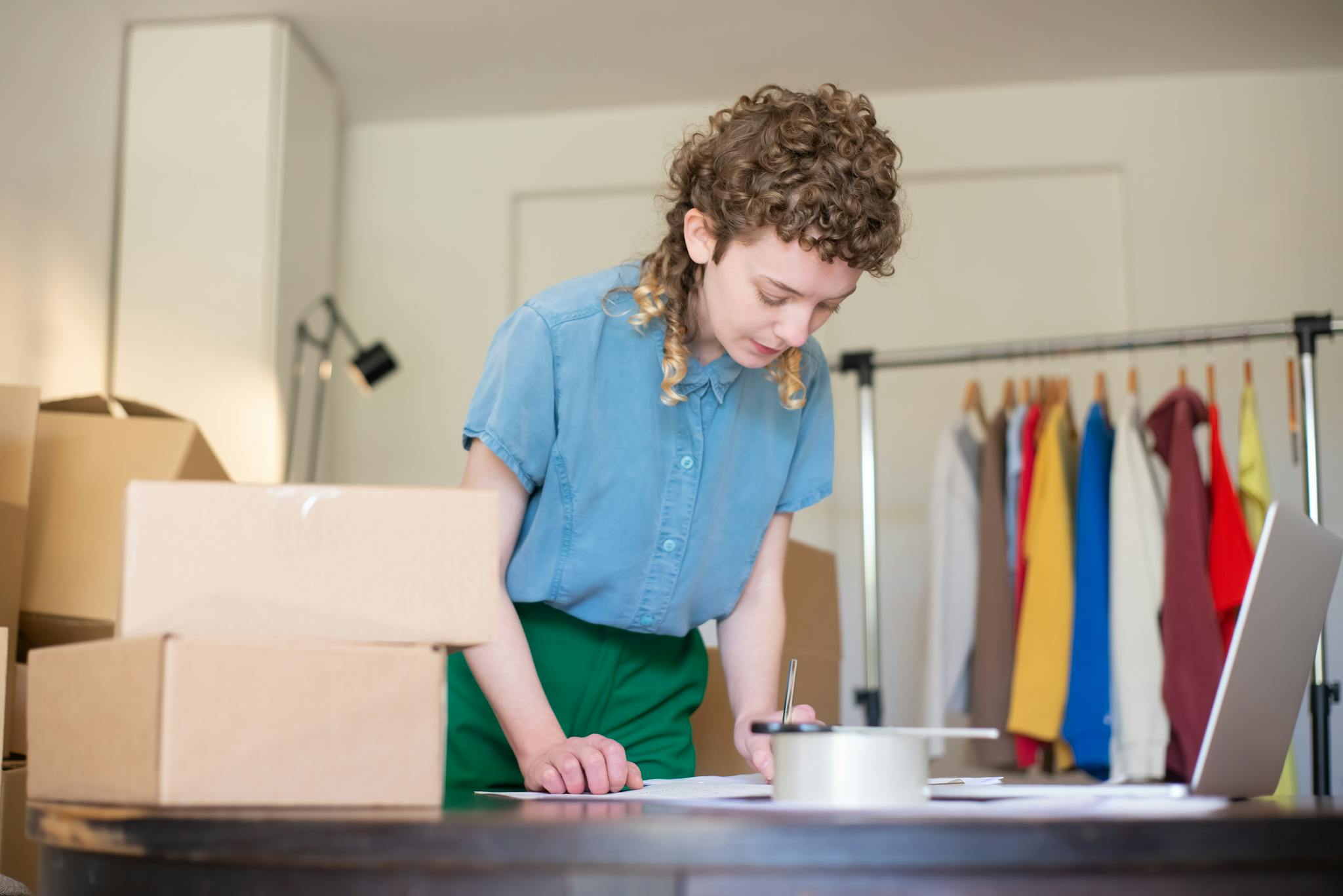 Woman packing online shop orders at home. Features boxes, clothing rack, and a laptop.